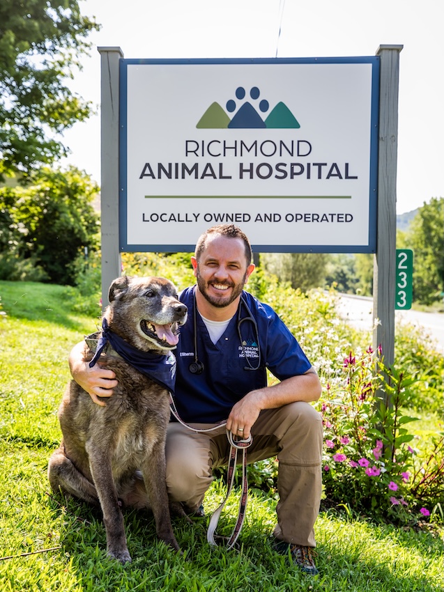 Dr. Nicholas Sherman kneeling with a senior dog beside the Richmond Animal Hospital sign in Richmond, VT